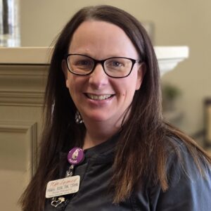 A person in medical scrubs with a smile, wearing glasses and a name tag.