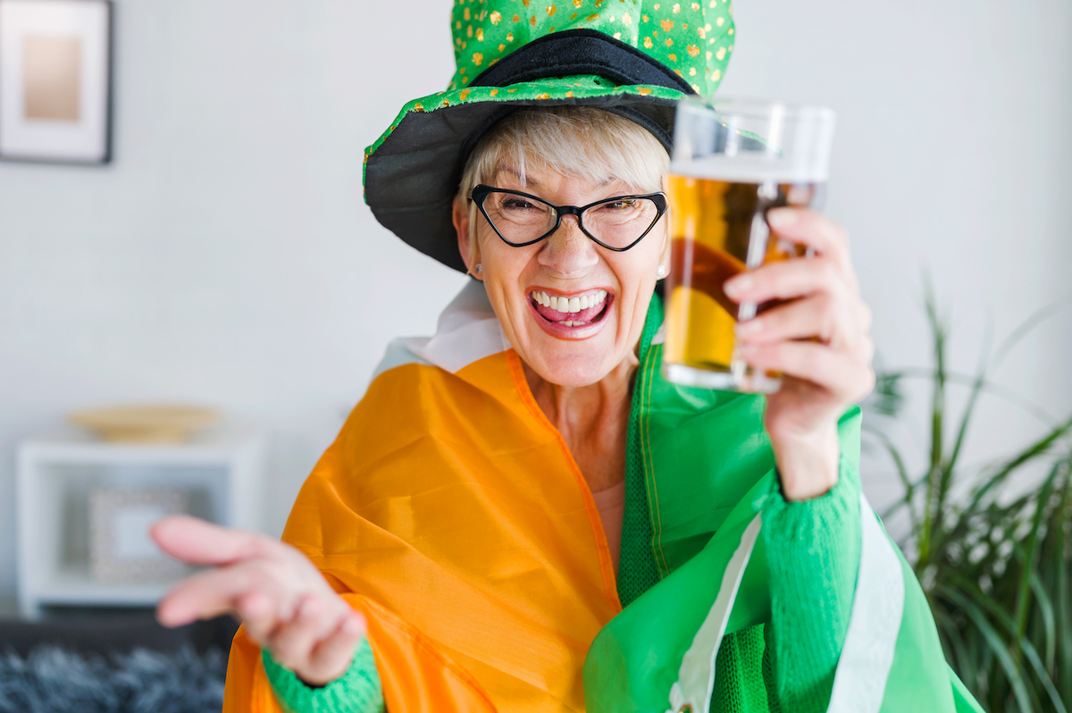 Person wearing a green hat and glasses smiles while holding a glass of beer. They are draped in an orange, white, and green flag.