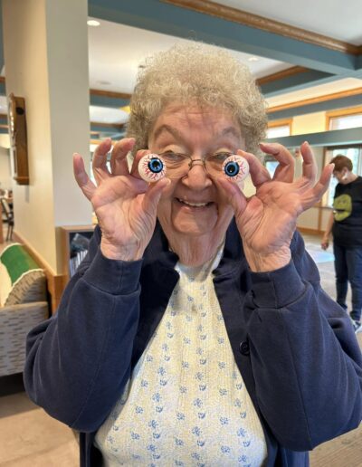 An older woman smiles and holds two fake eyeballs in front of her eyes inside a brightly lit room.