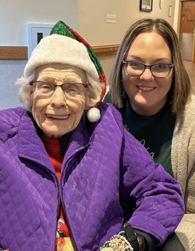 An elderly woman in a Santa hat and purple jacket sits next to a younger woman with glasses, both smiling at the camera indoors.