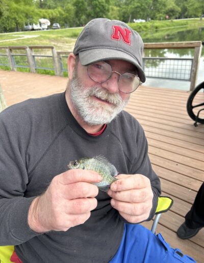 An older man with a gray beard and glasses holds a small fish while sitting on a dock by a pond. He wears a dark sweatshirt and a gray cap with a red "N" on it.
