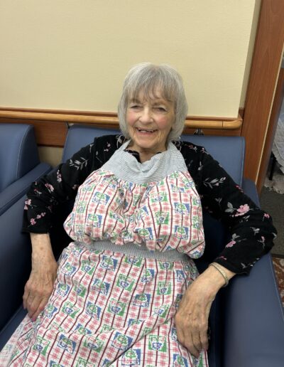 An elderly woman with gray hair sits in a blue chair, smiling, wearing a black floral top and a red, white, and blue checkered apron.