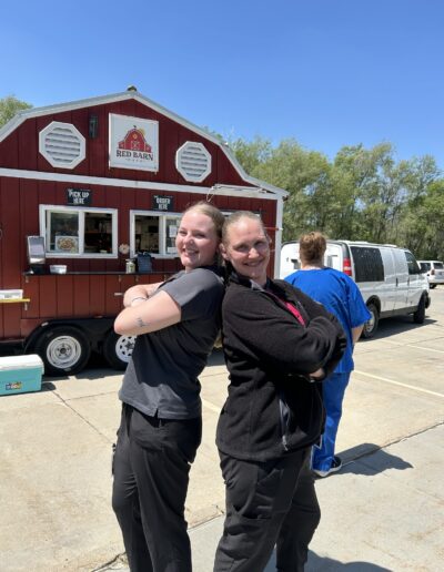 Two women in black outfits stand back-to-back and smile in front of a red food truck named "Red Barn." Another person in blue scrubs stands nearby, facing away. Cars are parked in the background.