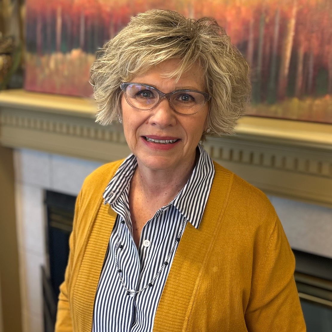 An older woman with short blond hair and glasses smiles at the camera, wearing a yellow cardigan and striped shirt, standing indoors in front of a fireplace and a colorful painting. An older woman with short blond hair and glasses smiles at the camera, wearing a yellow cardigan and striped shirt, standing indoors in front of a fireplace and a colorful painting.