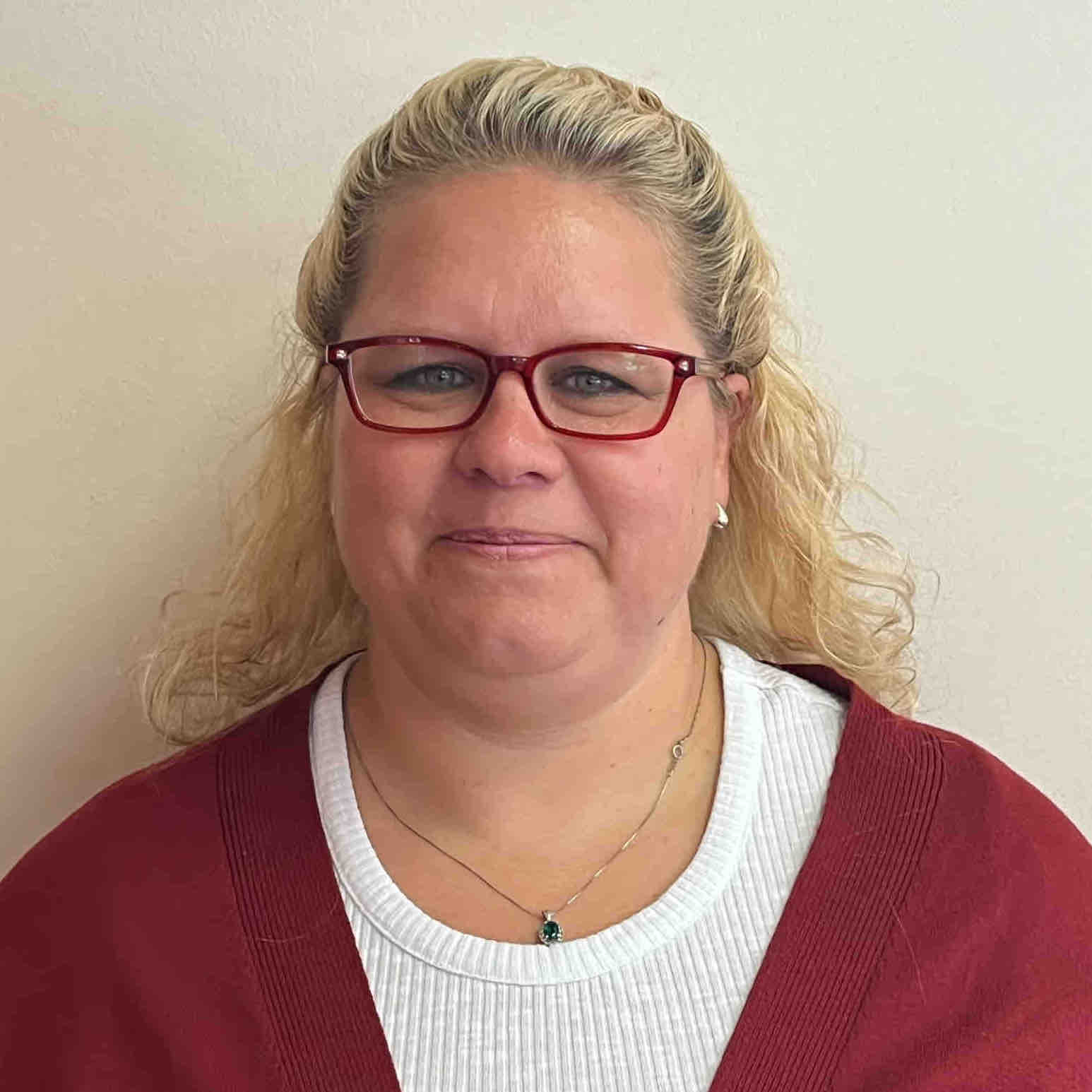 A woman with curly blonde hair, wearing red glasses, a white shirt, and a red cardigan, stands against a plain light-colored wall. A woman with curly blonde hair, wearing red glasses, a white shirt, and a red cardigan, stands against a plain light-colored wall.