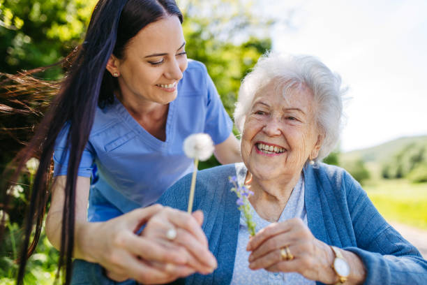 A young caregiver helps an elderly woman hold flowers while they both smile outdoors on a sunny day. A young caregiver helps an elderly woman hold flowers while they both smile outdoors on a sunny day.