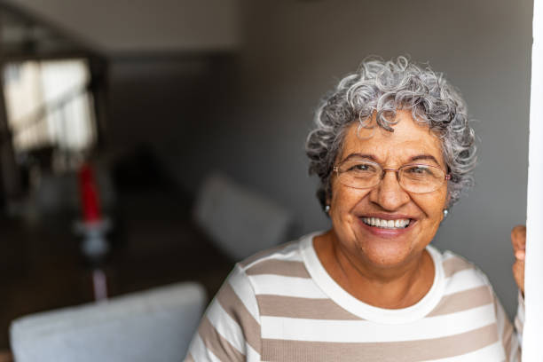 Older woman with curly gray hair and glasses, wearing a striped shirt, standing indoors and smiling at the camera. Older woman with curly gray hair and glasses, wearing a striped shirt, standing indoors and smiling at the camera.