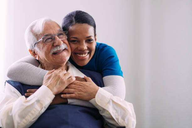 A young woman in scrubs hugs an older man with white hair and glasses from behind. Both are smiling in a bright indoor setting. A young woman in scrubs hugs an older man with white hair and glasses from behind. Both are smiling in a bright indoor setting.