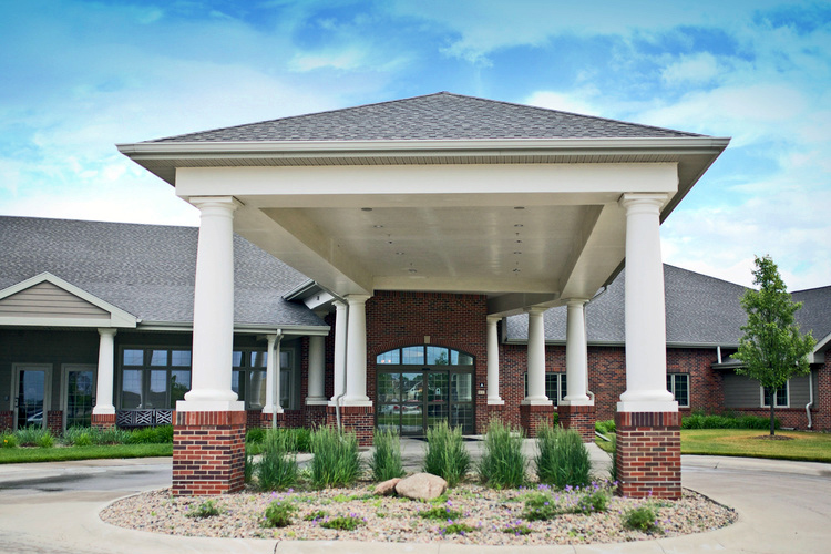 A single-story brick building with a covered entrance supported by white columns and a landscaped roundabout in front.