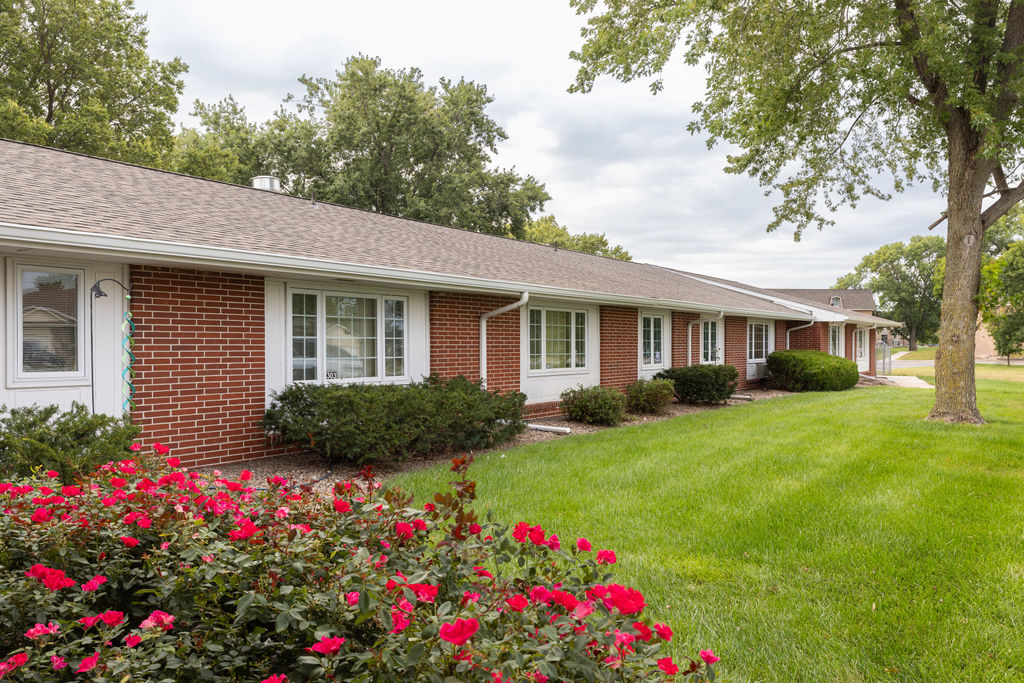 Single-story brick apartment building with white trim, surrounded by green grass, bushes, and blooming red flowers under a cloudy sky.