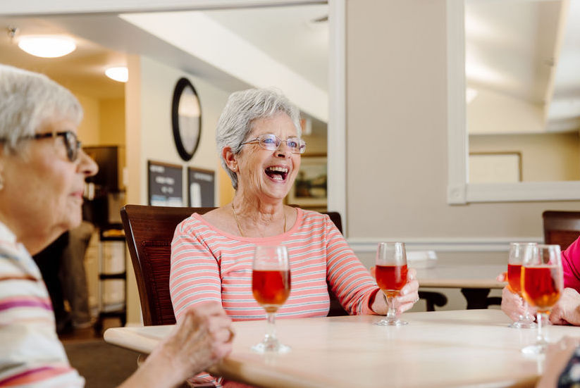 Three older women sit at a table in a brightly lit room, holding glasses of a red beverage and smiling while having a conversation.