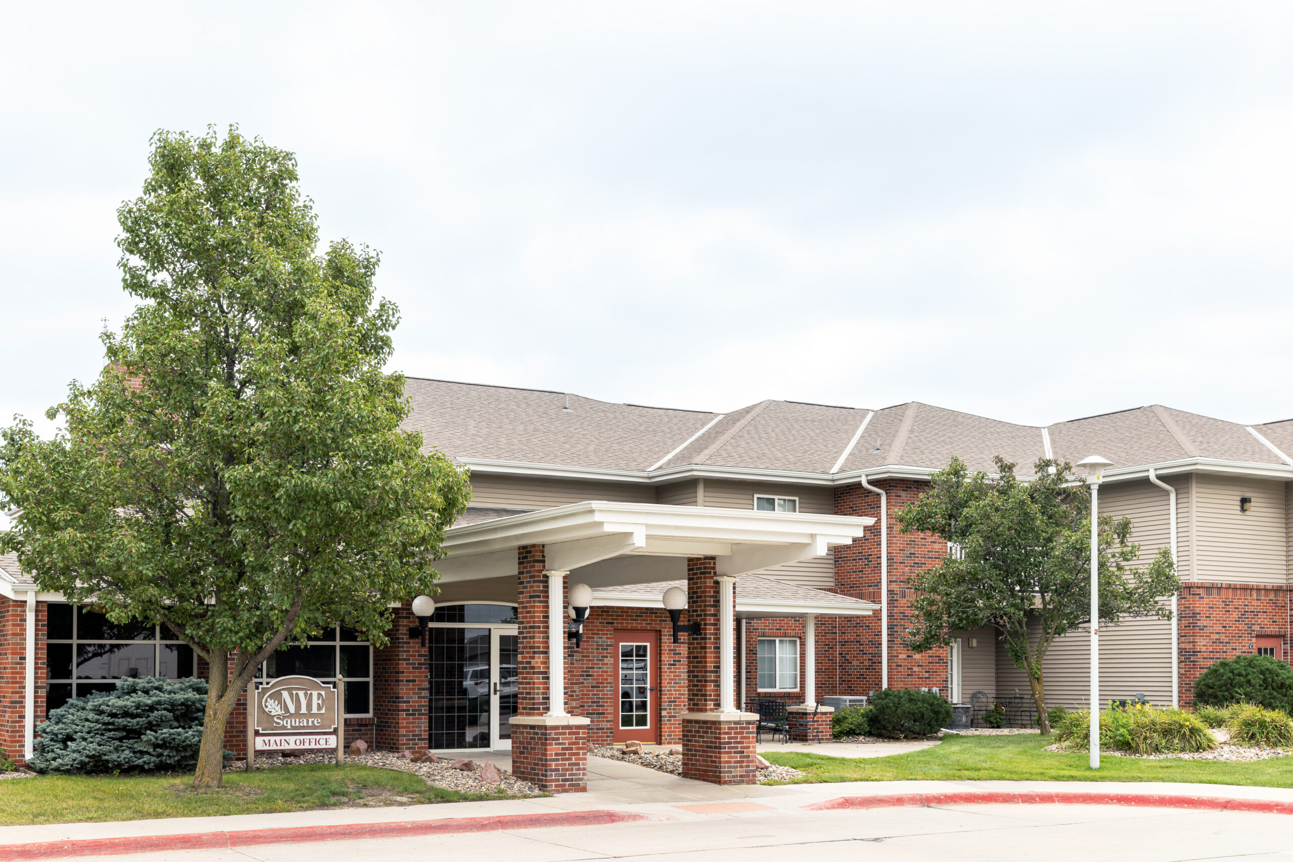 Exterior of a modern brick building with a covered entrance and white columns, featuring a sign that reads "Vive Senior Living Main Office" near a tree and landscaped bushes. Exterior of a modern brick building with a covered entrance and white columns, featuring a sign that reads "Vive Senior Living Main Office" near a tree and landscaped bushes.