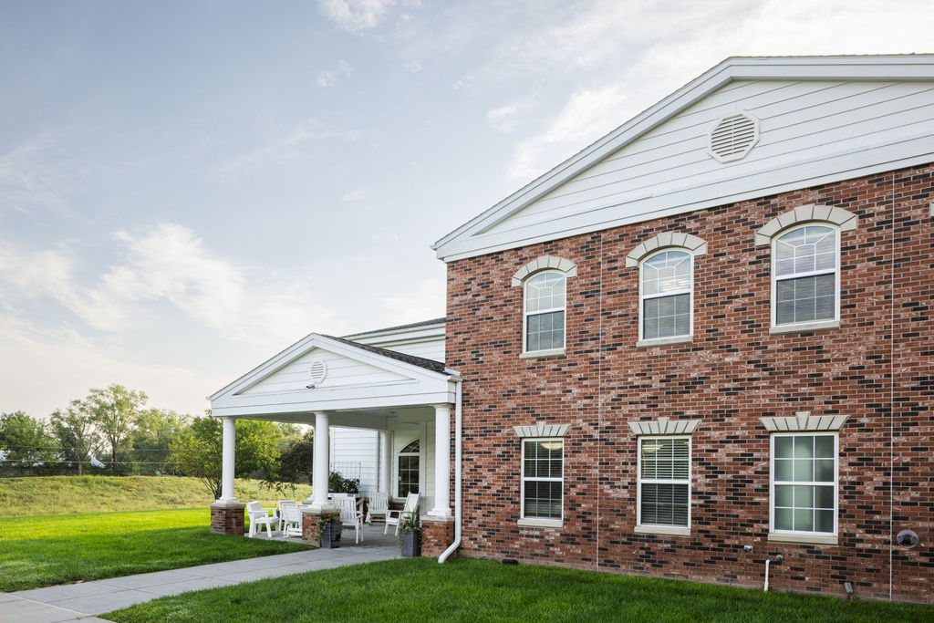 A two-story brick building with white trim, arched windows, and a covered front porch with chairs, set on a grassy lawn under a partly cloudy sky. A two-story brick building with white trim, arched windows, and a covered front porch with chairs, set on a grassy lawn under a partly cloudy sky.