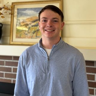 A young man in a light gray quarter-zip pullover stands indoors in front of a brick fireplace and a framed landscape painting.
